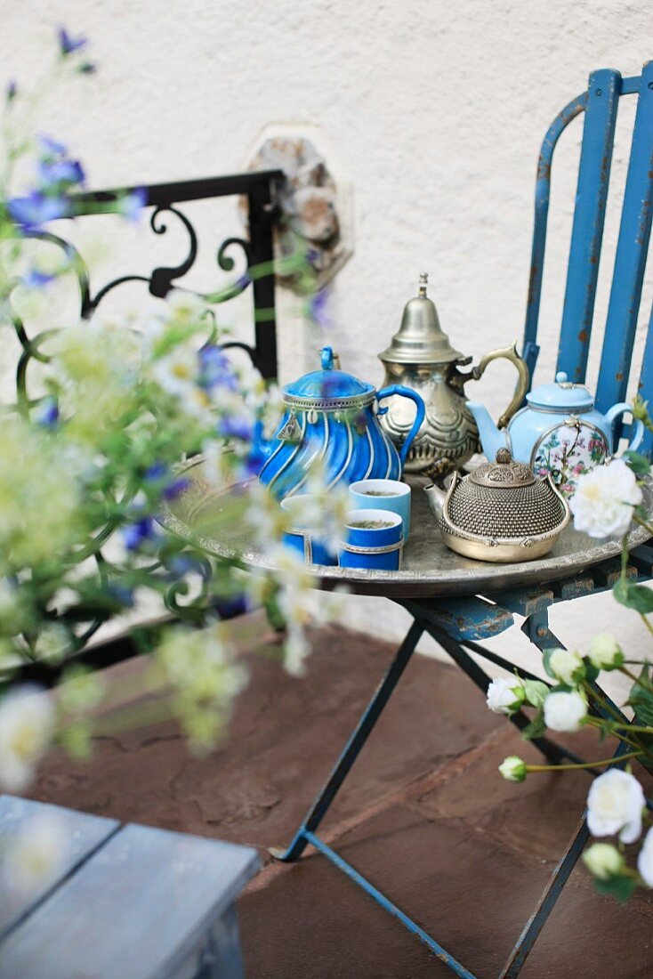 Silbertablett mit ausgefallenen, antiken Teekannen auf blau lackiertem Gartenstuhl; Terrasse mit floralem Brüstungsgeländer