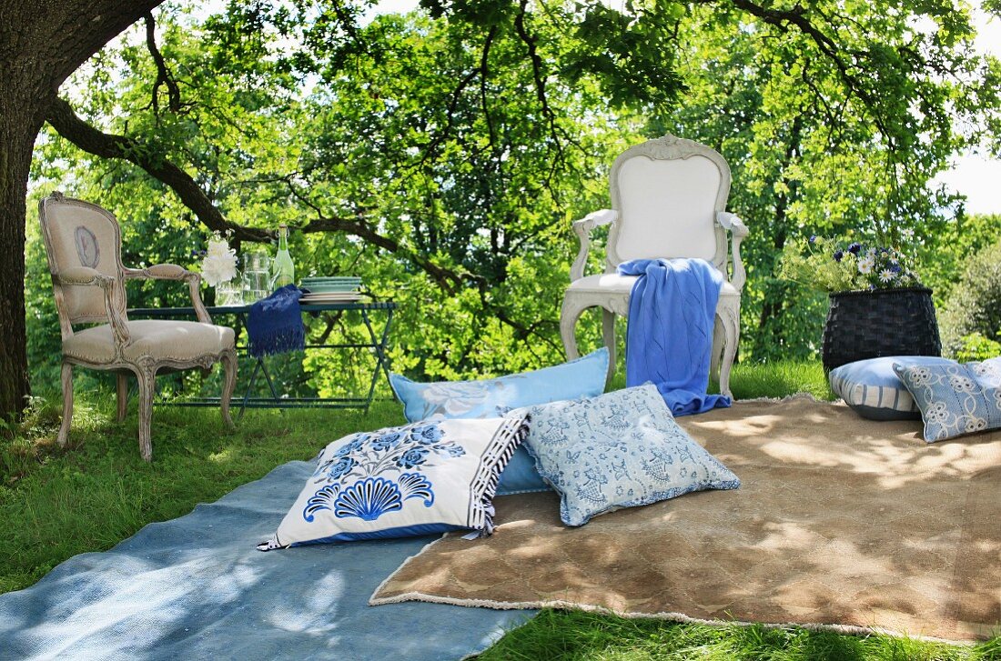 Stilvolle, in zartem Blau-Beige gehaltene Picknick-Zone unter Gartenbäumen, mit einladenden Kissen, Decken und Rokokostühlen neben einem Gartentisch