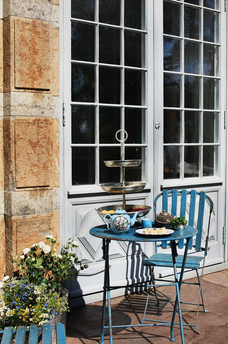 Vintage garden furniture made from blue-painted metal in front of terrace door of old house - afternoon tea with silver cake stand and blue china teapot