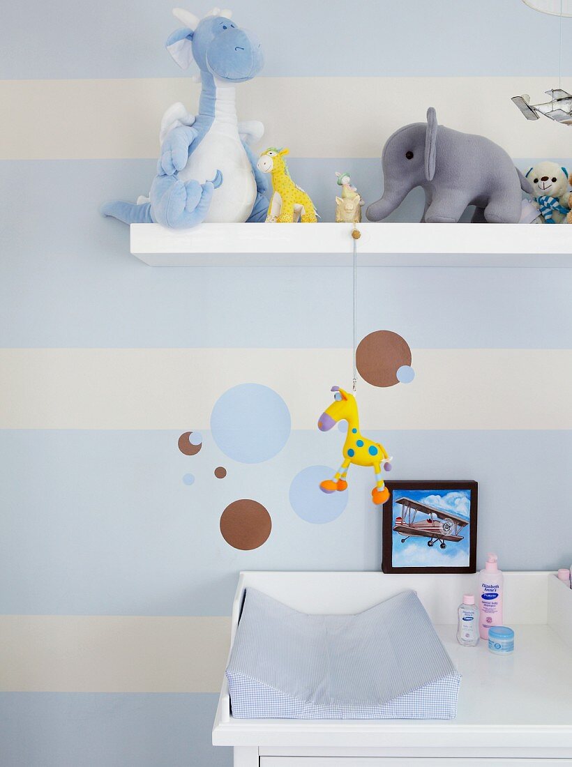 Mit hellblauen Streifen und Kreisen gestaltete Kinderzimmerwand; auf dem Wandboard verschiedene Plüschtiere und auf der weißen Wickelkommode eine hellblaue Wickelauflage