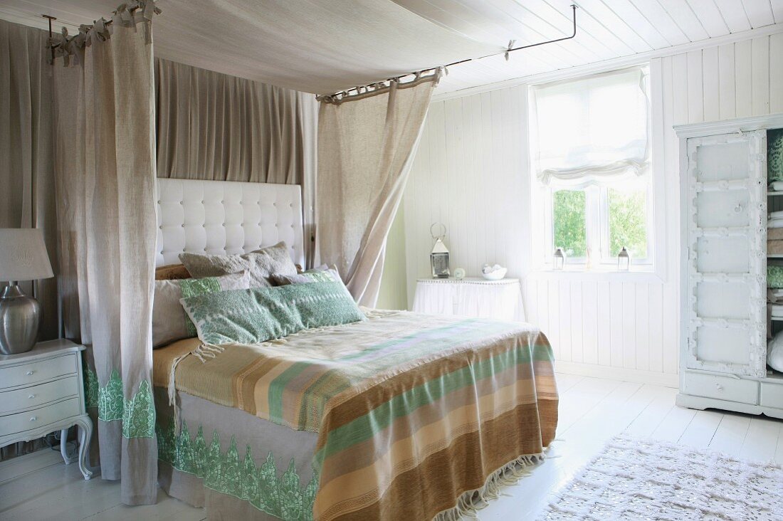 Feudales Bett mit gestepptem Kopfende und luftigen, verzierten Bettvorhängen in sonnigem Schlafzimmer