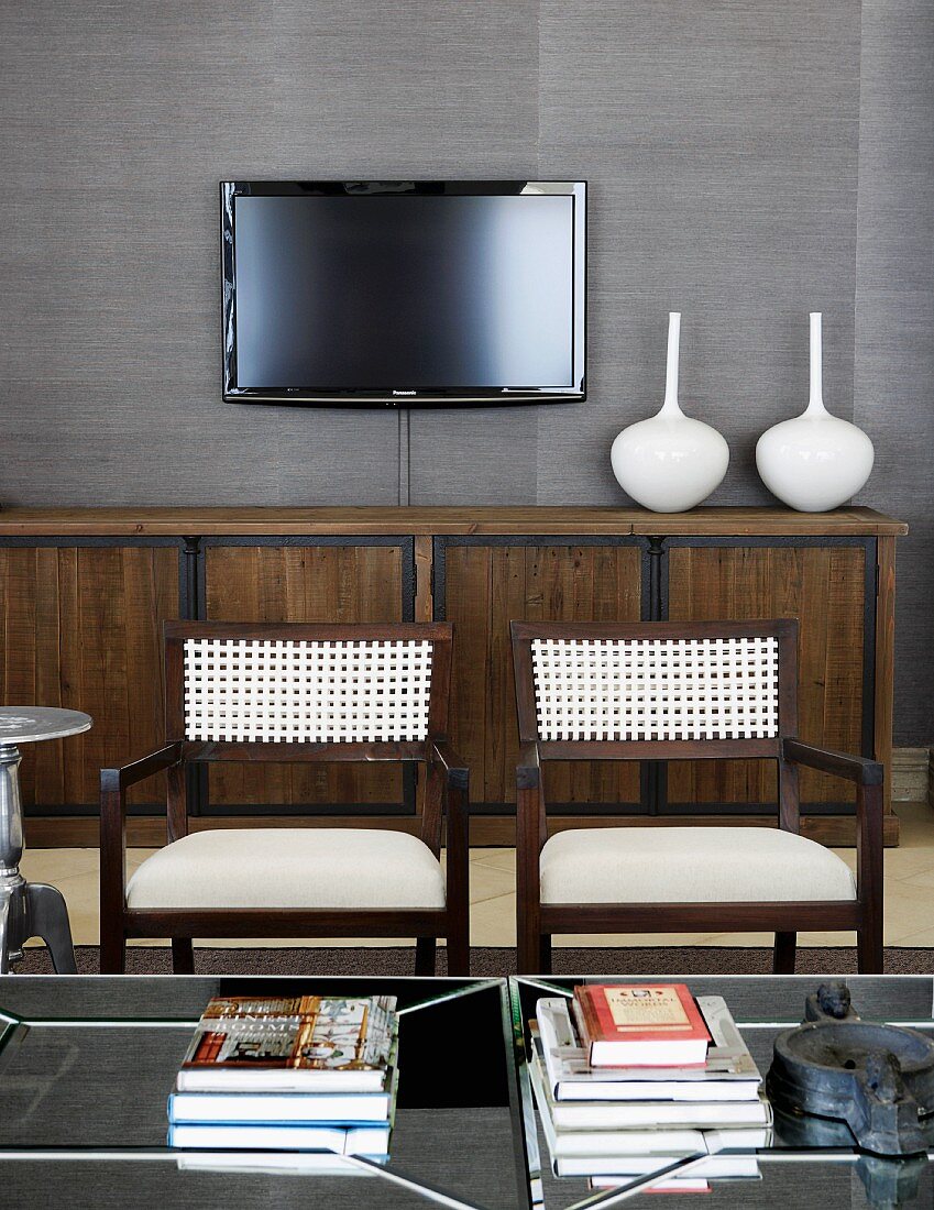Braun-schwarzes Sideboard mit weissen Vasen und Flachbildfernseher an grau tapezierter Wand; im Vordergrund zwei elegante Armstühle mit geflochtener Stuhllehne