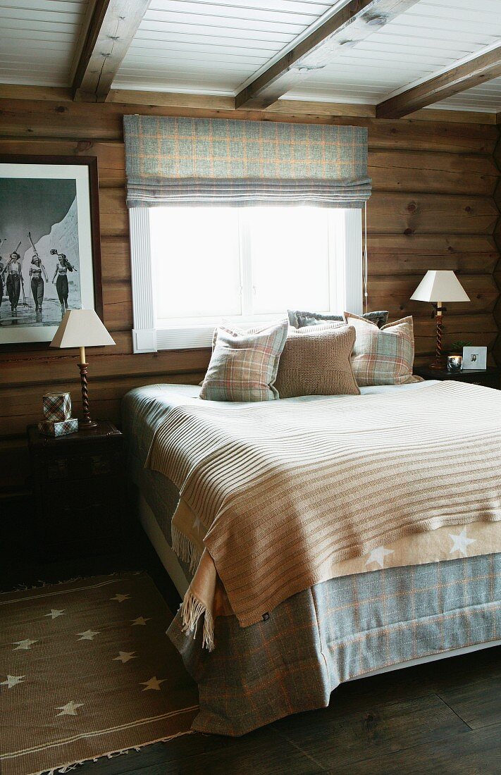 Double bed with bedspread and scatter cushions below window in rustic bedroom in wooden house