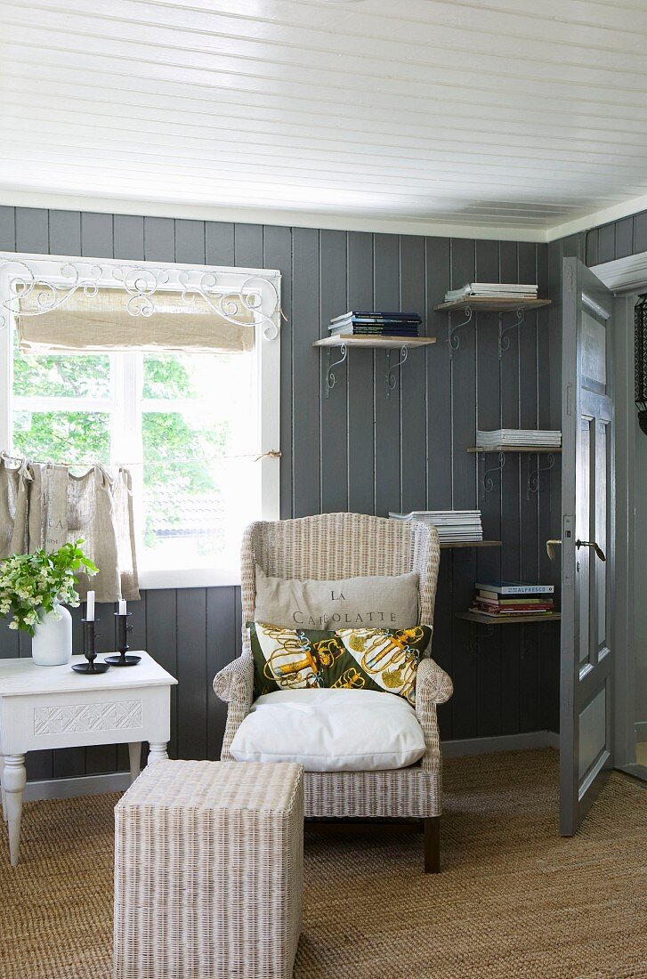 Comfortable wicker reading chair and stool in front of sunny window and wood-panelled wall with small bookshelves