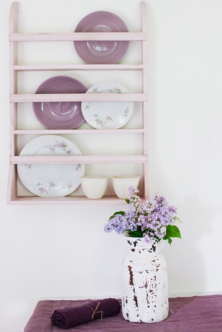 Geschirrregal in Zartrosa an weisser Wand; auf dem Tisch eine Vase im Shabby Stil mit duftendem Flieder