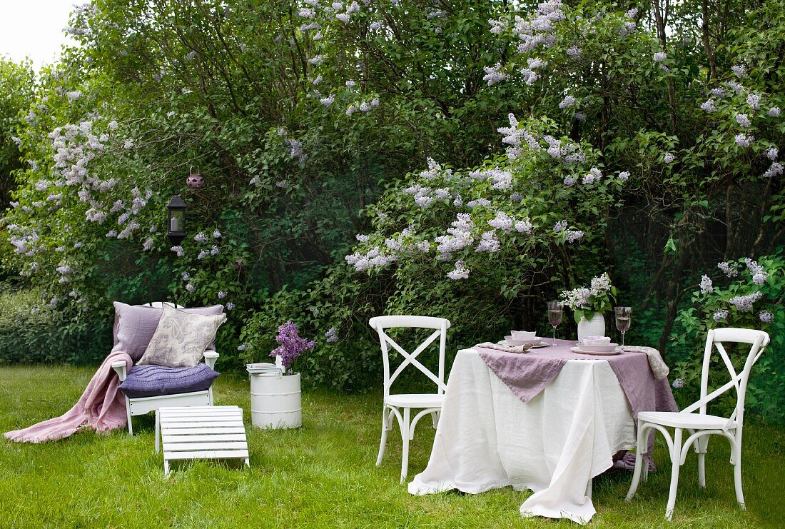 Idyllischer Garten mit blühendem Flieder; im Vordergrund ein gedeckter Tisch und ein Gartenstuhl mit bequemen Kissen