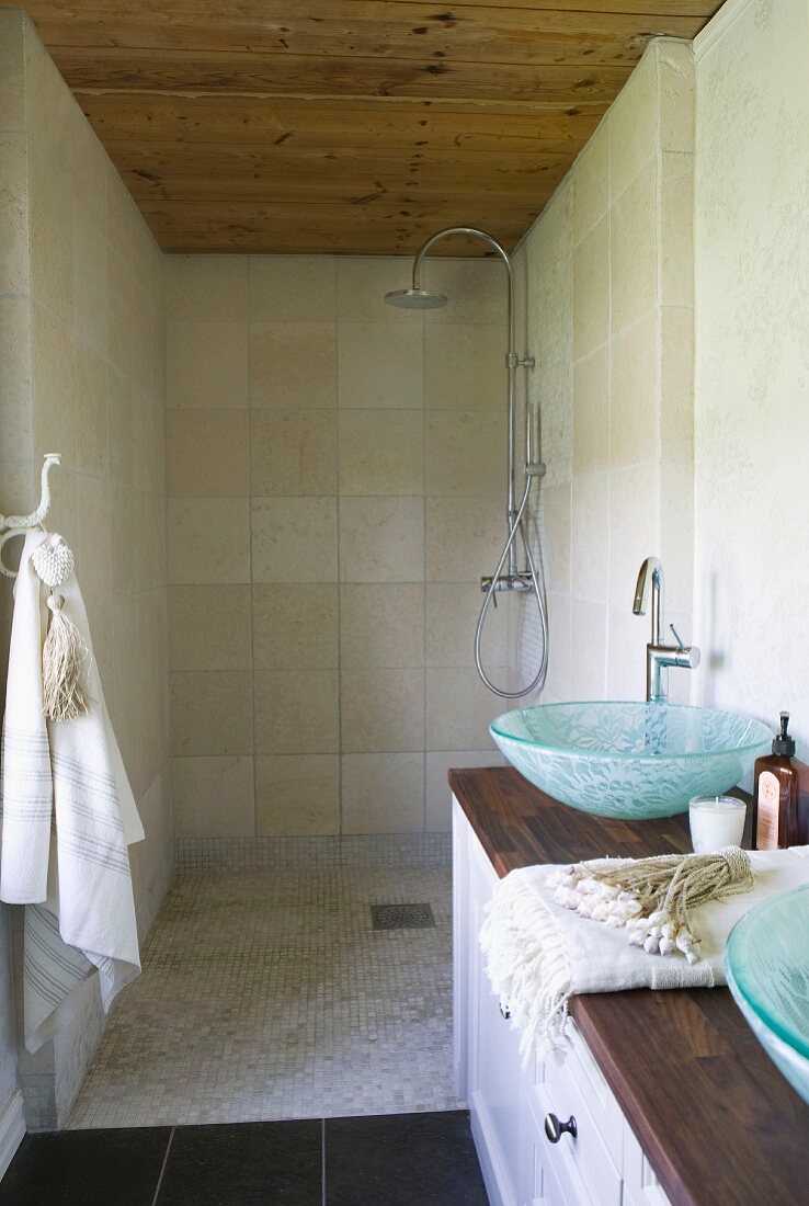 Duschbrause in schmalem Duschraum mit Holzdecke; im Vordergrund ein langer Holzwaschtisch mit zwei gläsernen Waschschüsseln