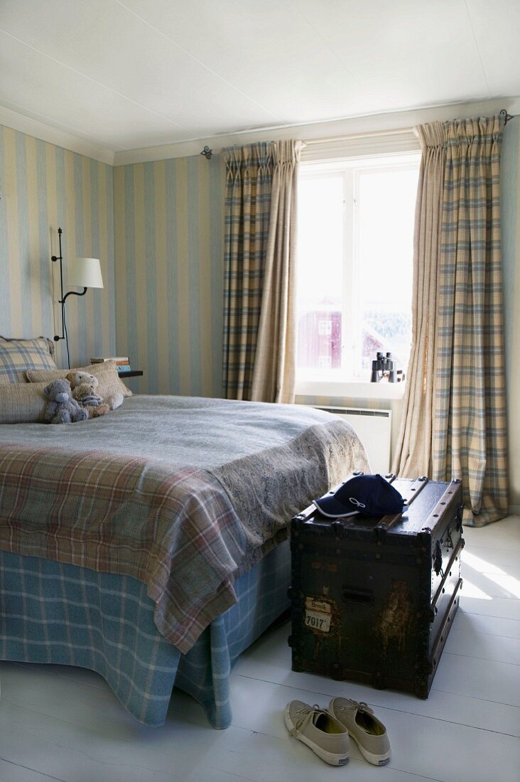 Summery bedroom in subtle colours with pale striped wallpaper and checked curtains and bed linen; antique wooden trunk at foot of bed