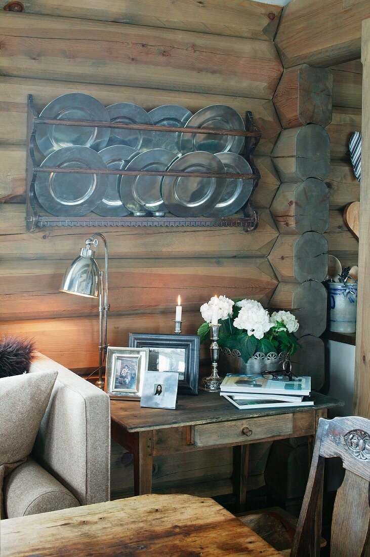 Gerahmte Photos und Retro Tischleuchte auf rustikalem Tisch vor Holzwand mit Zinnteller Sammlung auf gehängtem Bord