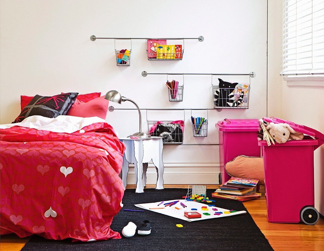Girl's bedroom with red bed linen with heart motif; pink bins and wire baskets on wall for storing toys
