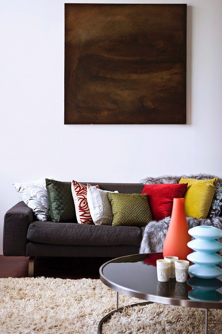 Düsteres Gemälde über Sofa mit farbenfrohen Dekokissen; originelle Vasen auf modernem Stahlglas-Tisch