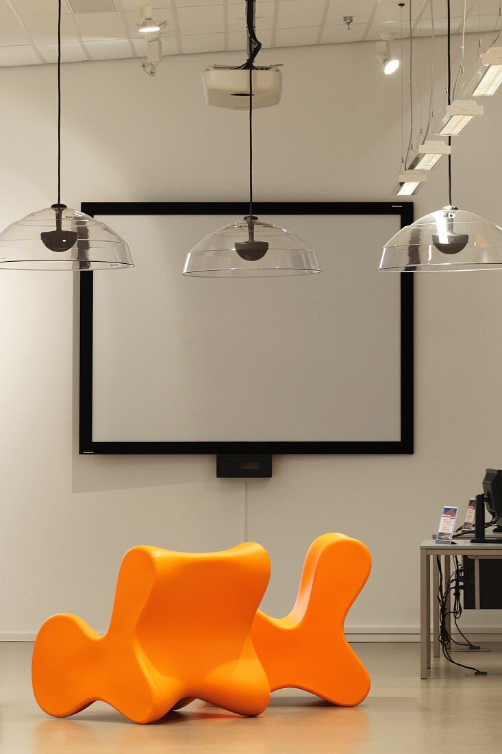Leuchtend orange Stühle und transparente Hängeleuchten vor einer Leinwand mit schwarzer Rahmung (Bibliothek Hoofddorp-Centrale)