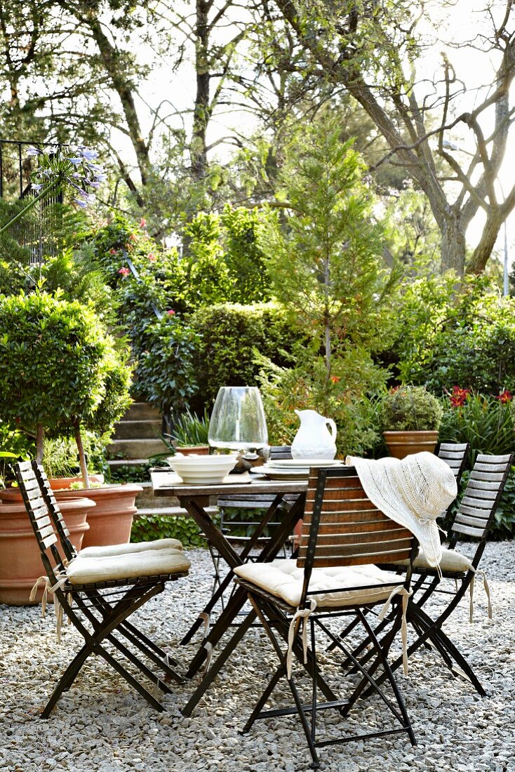 Classic garden table and chairs on gravel floor; groups of shrubs, trees and box bushes in terracotta pots in background