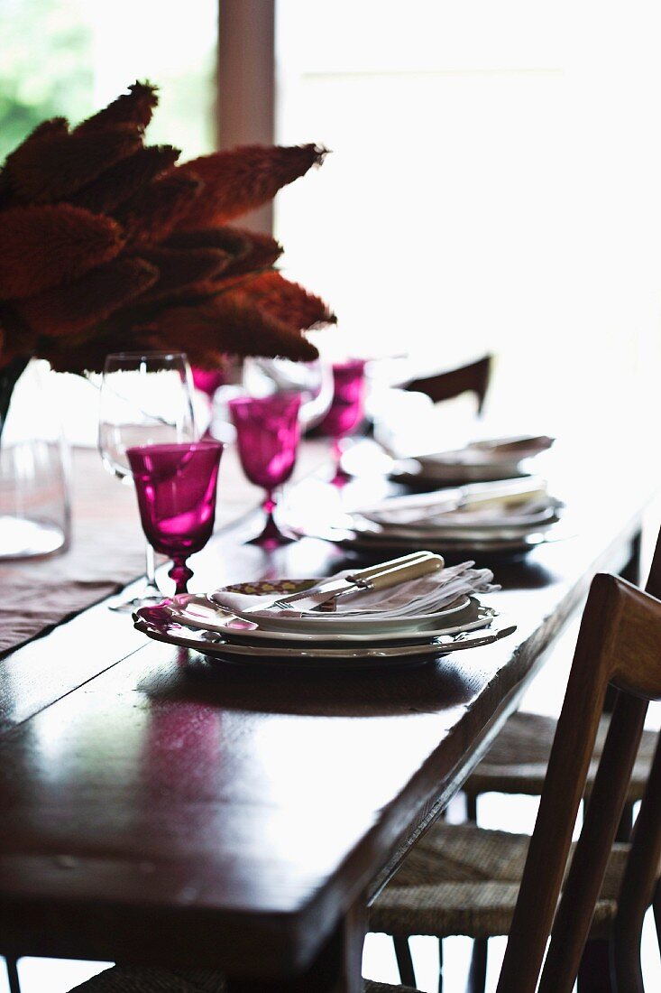 Magentafarbene Weingläser und exotischer Tischschmuck auf festlich gedecktem, antikem Holztisch mit einfachen Geflechtstühlen