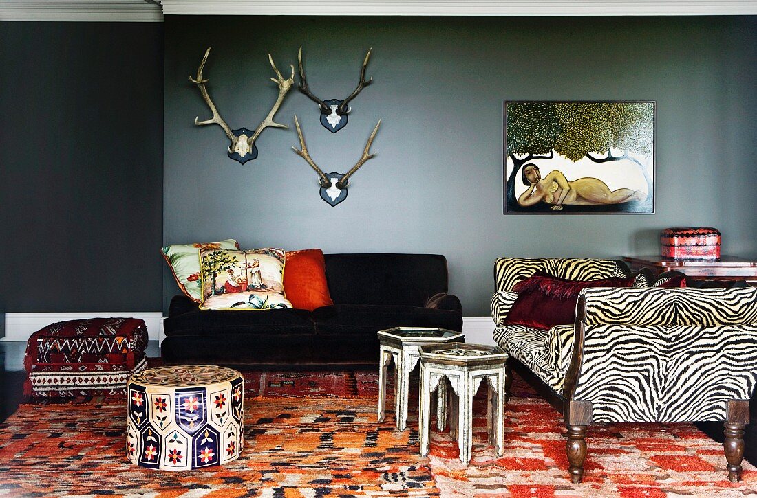 Stags' antlers and naïve artwork above eclectic seating in wild mixture of styles - antique sofa with zebra patterned upholstery combined with Oriental side tables and pouffes
