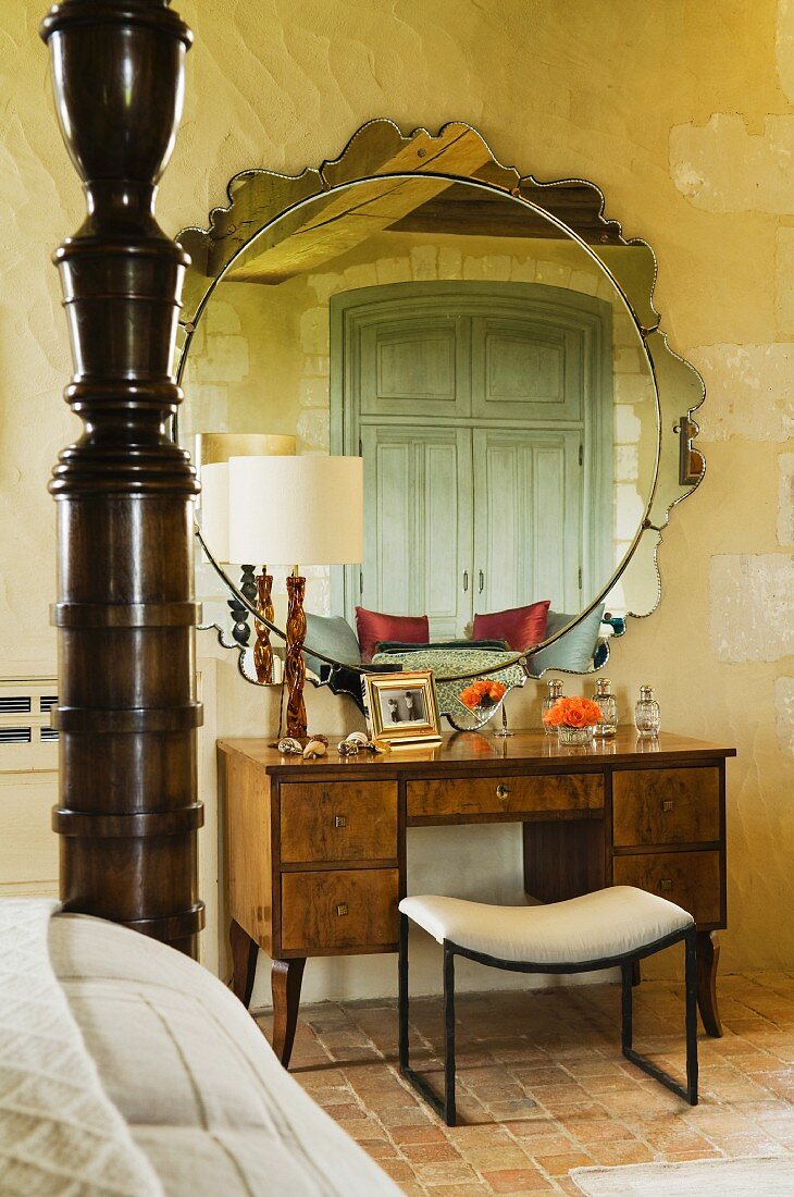 Frisiertisch und Hocker vor rundem Spiegel mit Verzierung und teilweise sichtbares Bett mit Holzsäule