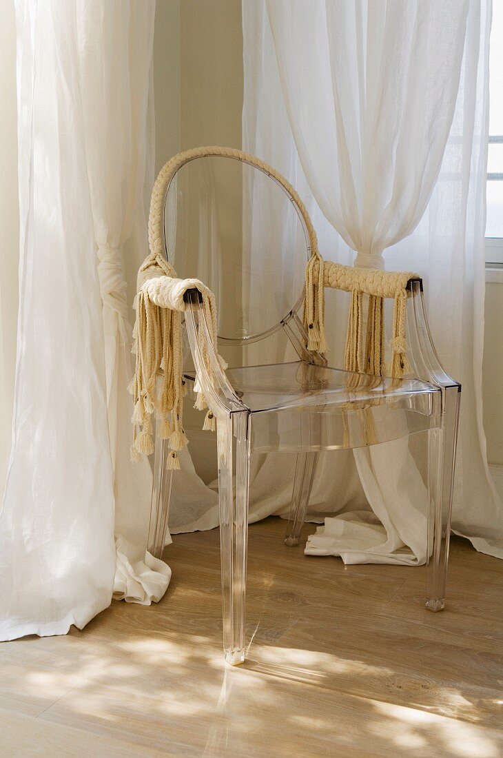 Decorated, plastic ghost chair in corner of bedroom in front of white, translucent curtains
