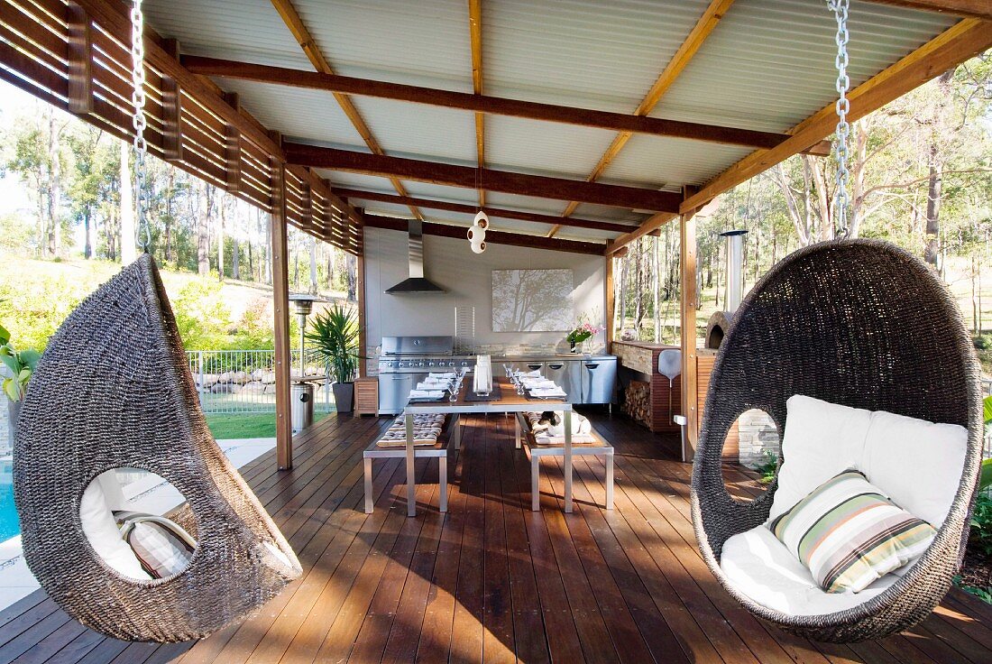 Roofed outdoor kitchen next to pool with long dining table and wicker hanging chairs