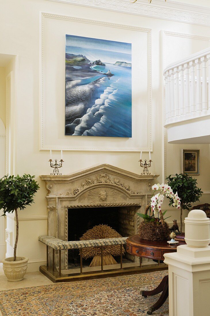 Open fireplace with stone mantelpiece below modern artwork on white, wood-panelled wall in foyer with partially visible gallery