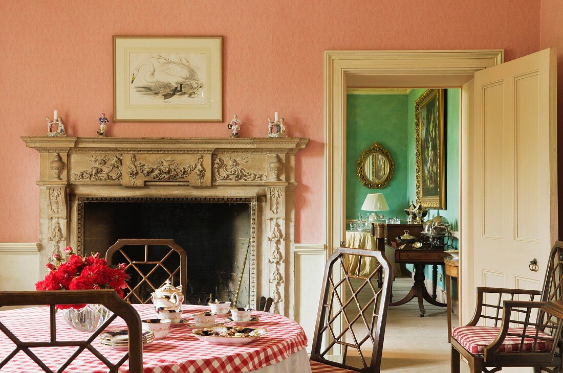 Rosafarbenes Frühstückszimmer mit ländlicher Tischdecke auf Tisch vor offenem Kamin mit Steinschürze und Blick durch offene Tür ins Nebenzimmer