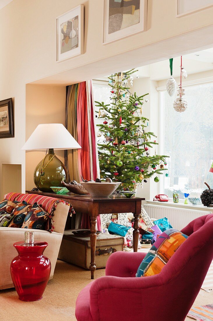 Geschmückter Weihnachtsbaum und Geschenkpäckchen in Fenstererker; pinkfarbener Sessel und roter Glasballontisch im Vordergrund