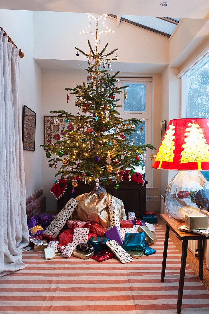 Traditionally decorated Christmas tree raised above mountain of presents in large bay window; lamp with glass balloon base and festive lampshade to one side