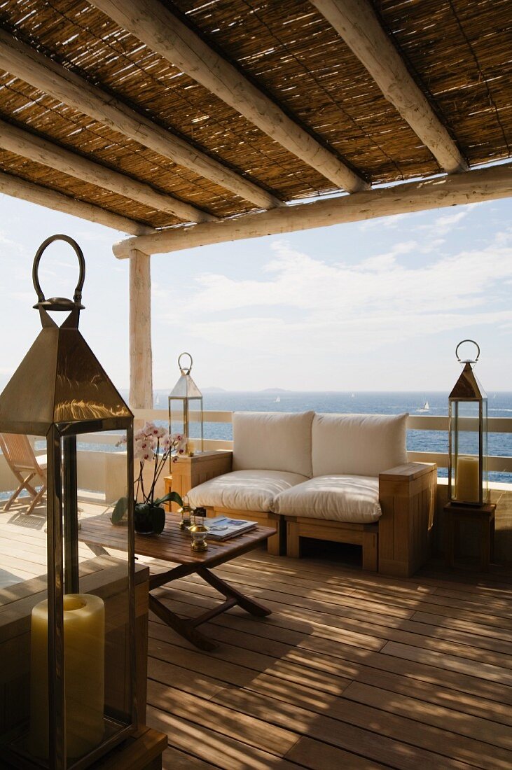 Sitzbank mit weissen Polstern zwischen Laternen auf Terrasse mit Strohdach und Blick auf das Meer