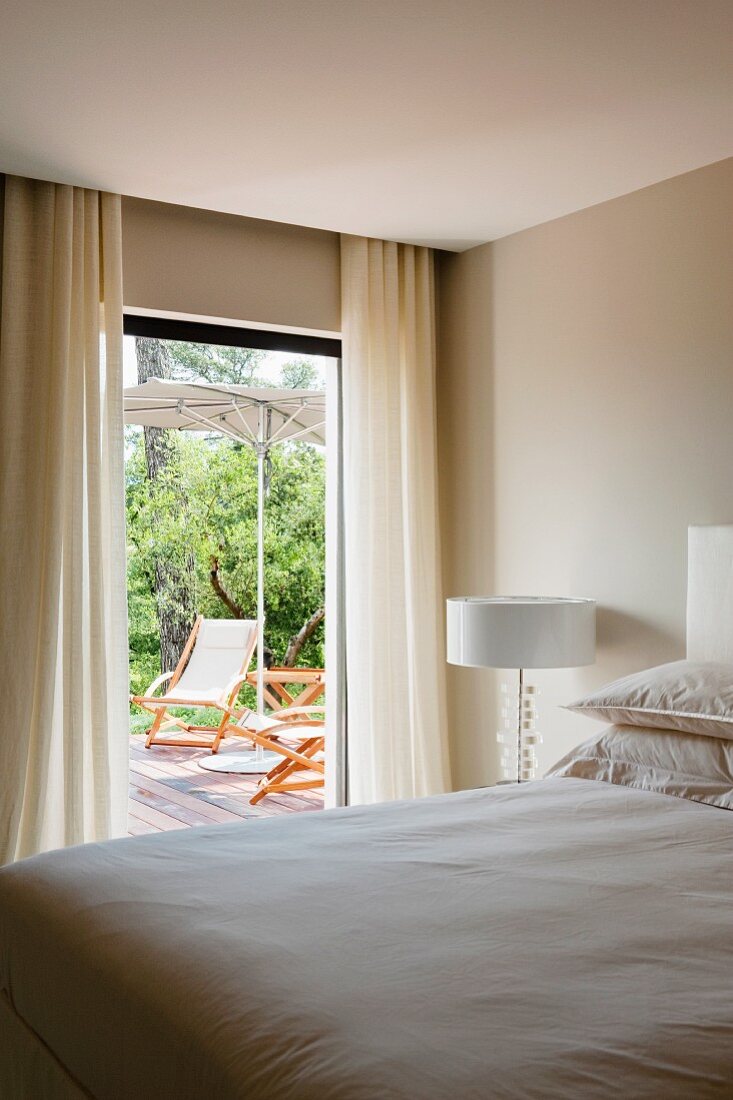 Blick über Doppelbett auf Terrassentür und Liegestühle unter Sonnenschirm