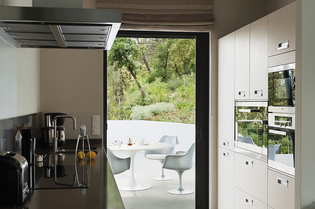 Designer Küche mit Geräten im Einbauschrank und Blick durch offene Terrassentür auf weiße Kunststoffstühlen aus Bauhauszeit