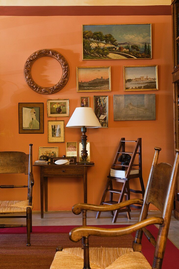 Antike Binsen-Armstühle vor apricotfarbener Wand mit einer Sammlung kleiner Landschaftbilder und antikem Wandtisch