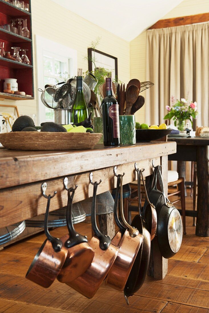 Massiver Holztisch im Essbereich auf Landhausdielenboden mit seitlich aufgehängten Kupferpfännchen, gedeckt mit Korbschale, Weinflaschen und Aufbewahrungsgefäßen