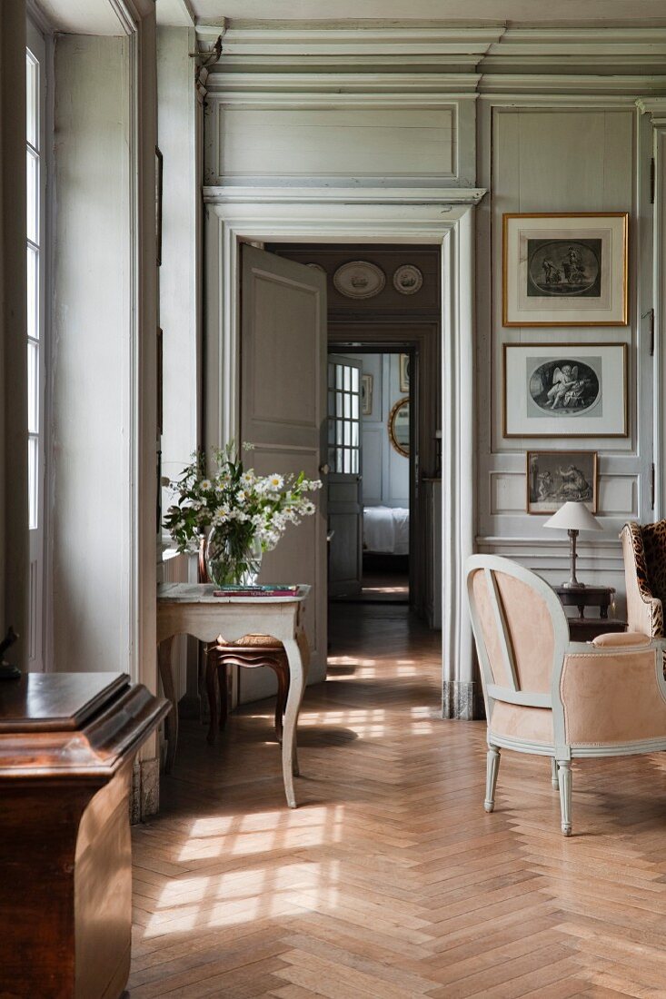 View through open interior doors of grand salon with antique side table below window and delicate armchair on herringbone parquet floor