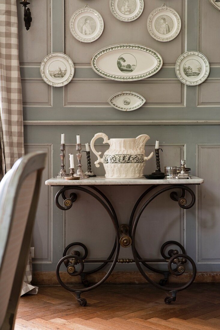Candlesticks and antique jug on washstand with metal frame below plates and platters hung on grey-painted wooden wall