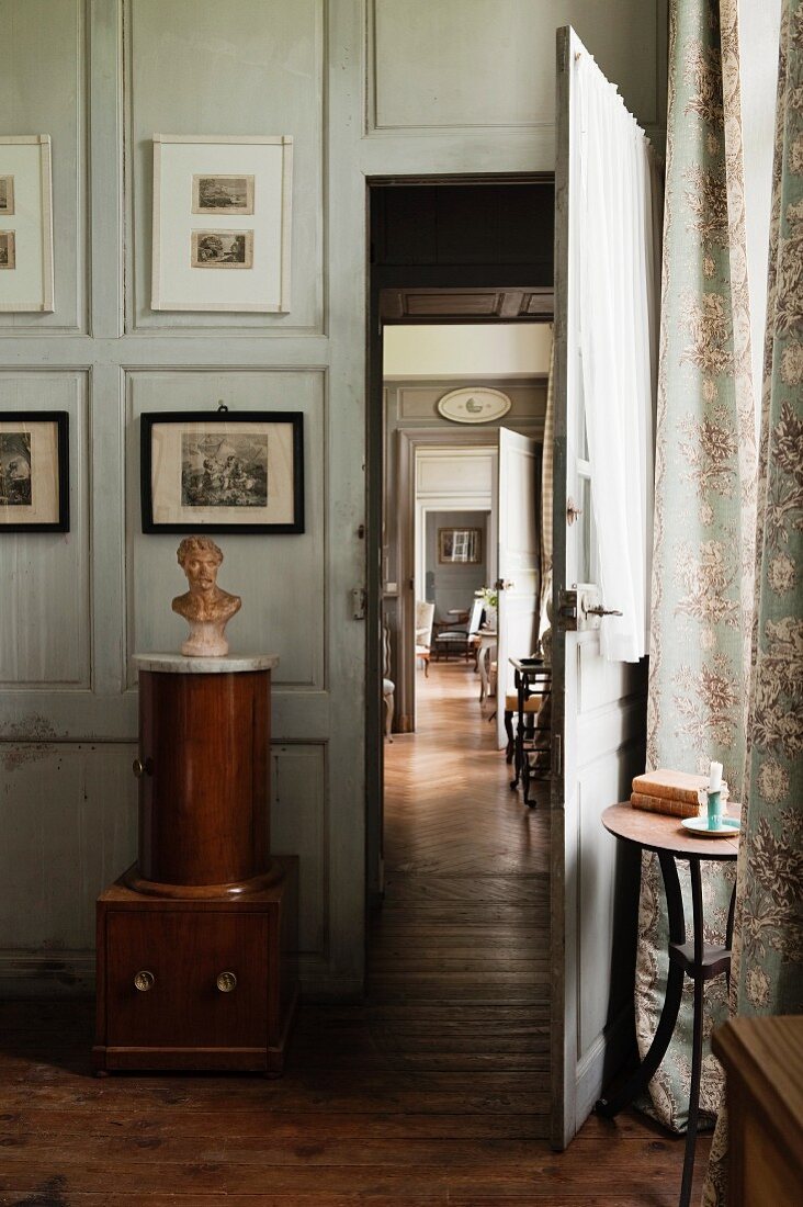 Kleinmöbel aus Holz mit Büste vor heller Holzpaneelwand neben Tür mit Blick durch offene Zimmertüren