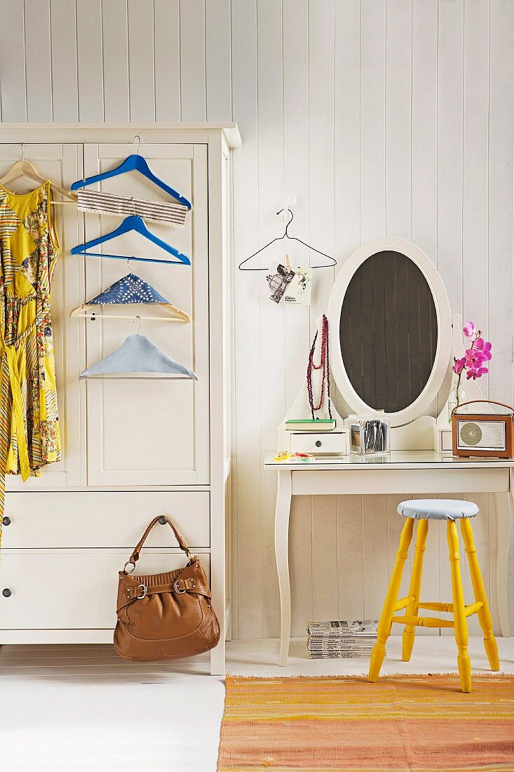 Dress and coathangers hanging on front of white-painted farmhouse wardrobe next to dressing table and yellow stool against white, wood-panelled wall