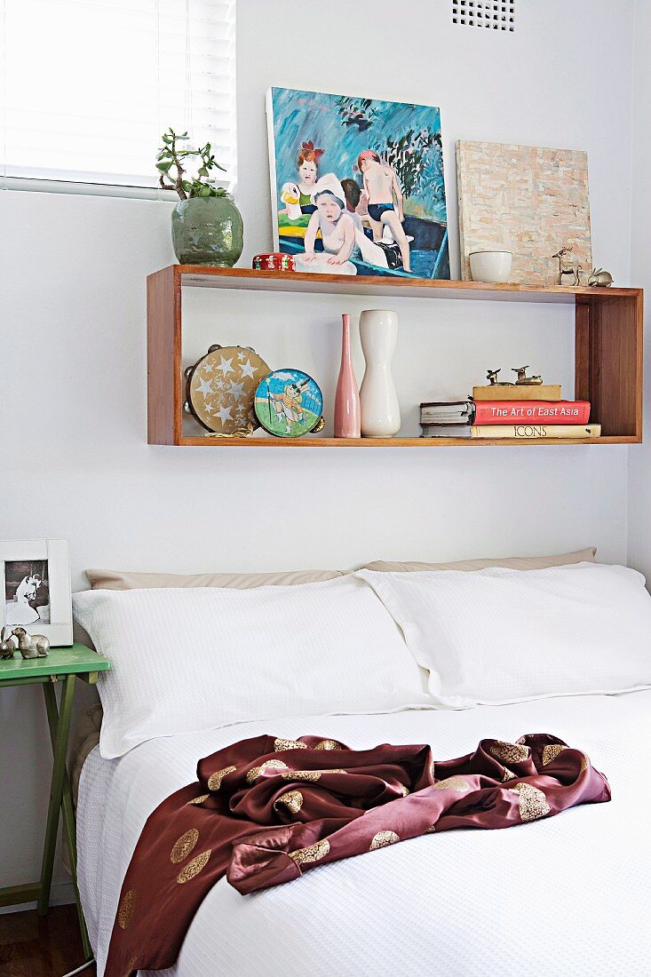Double bed with white bed linen below floating shelf of books and pictures