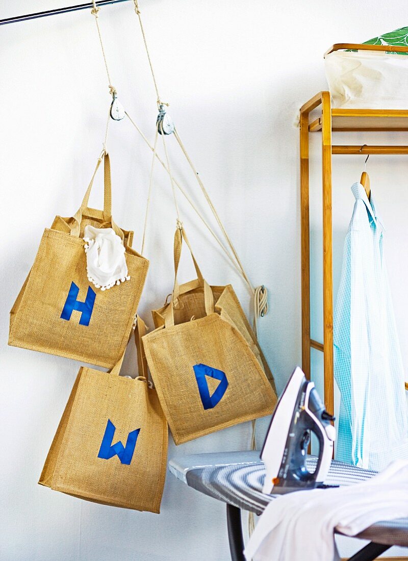 Aufgehängte Taschen aus Leinen mit blauen Buchstaben neben offener Garderobe und Bügeleisen auf Bügeltisch