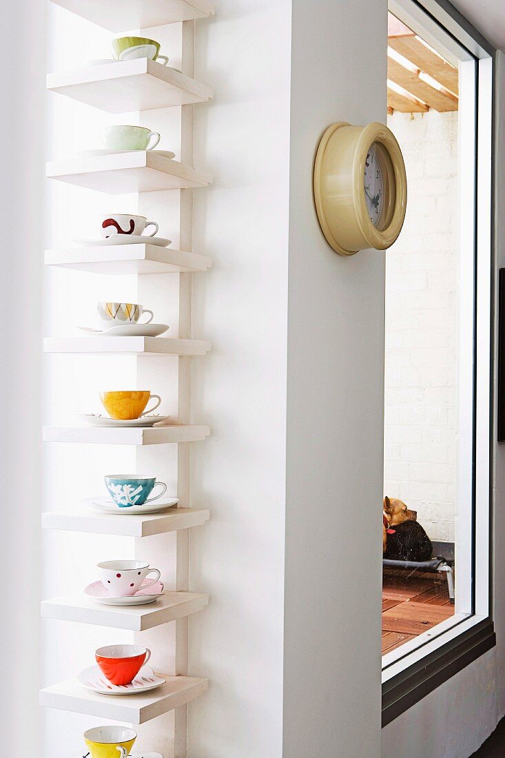 Coloured cups and saucers displayed on white shelves in window niche next to vintage submarine clock on wall