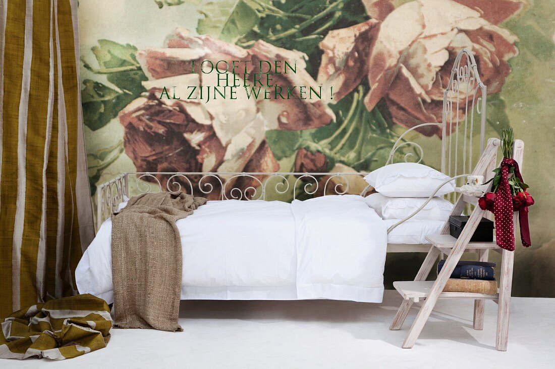 Verschnörkeltes Metallbett mit weißer Bettwäsche und Vintage Trittleiter vor Wand mit großflächigem Rosenmotiv