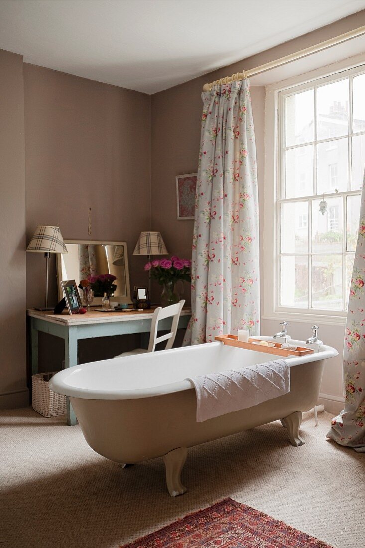 Free-standing vintage bathtub in front of window with floor-length curtains and dressing table with mirror in corner of room