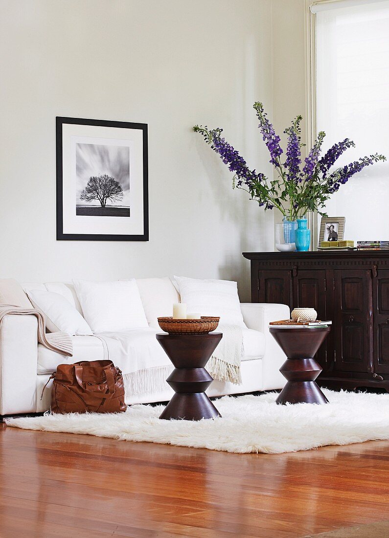 Elegant gehaltener Wohnbereich mit glänzendem Parkettfussboden und weißem Polstersofa an der Wand, davor dunkle trommelförmige Beistelltischchen, daneben dunkle Holzkommode mit Blumenstrauß in lila