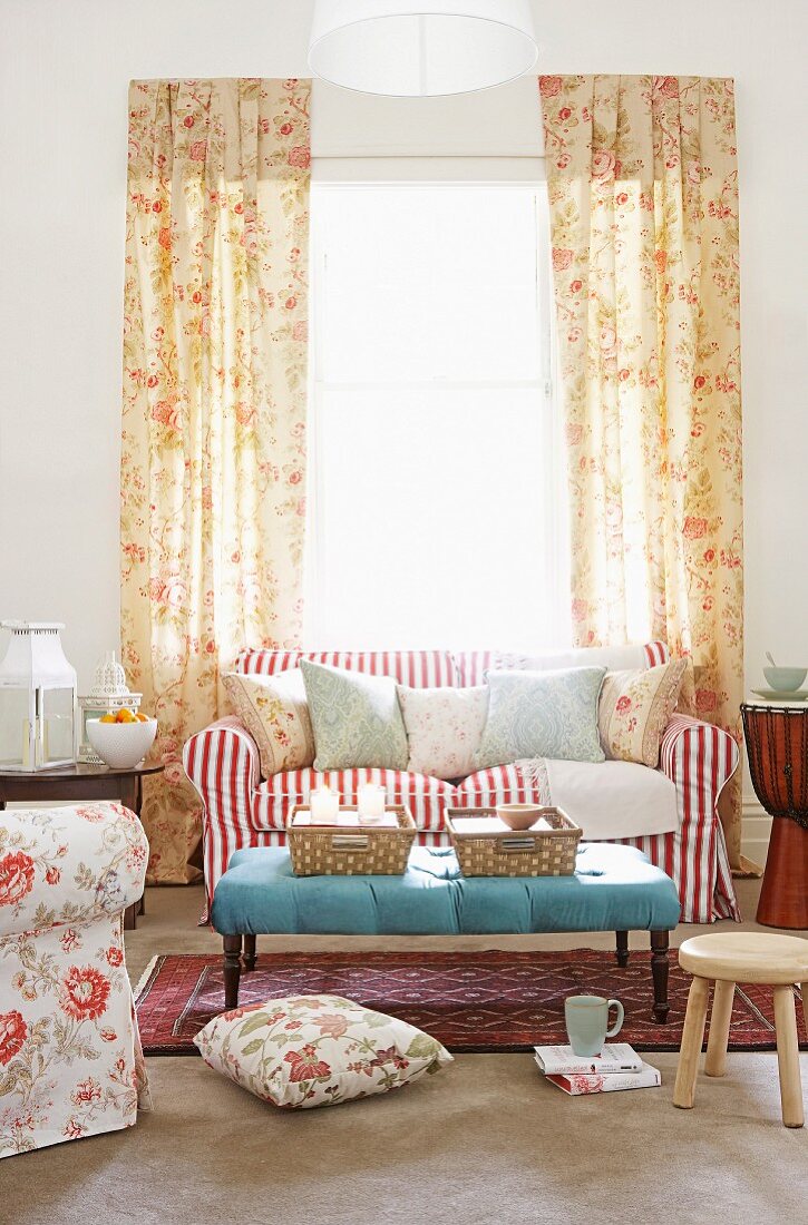 Gemütliches romantisches Wohnzimmer mit blumigen bodenlangen Vorhängen, vor dem hellen Fenster ein rot-weiß gestreiftes Polstersofa mit vielen Kissen und einem hellblau gepolsterten Couchtisch davor