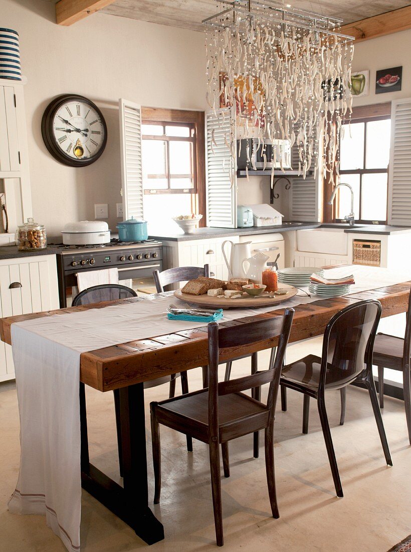 Rustikaler Esstisch aus Holz mit weißem Tischläufer und verschiedene Stühle unter maritimer Leuchte in ländlicher Küche