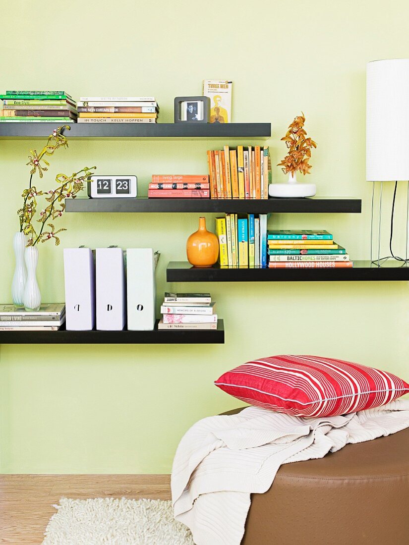 Books and box files on black floating shelves on pastel green wall and cushion on ottoman