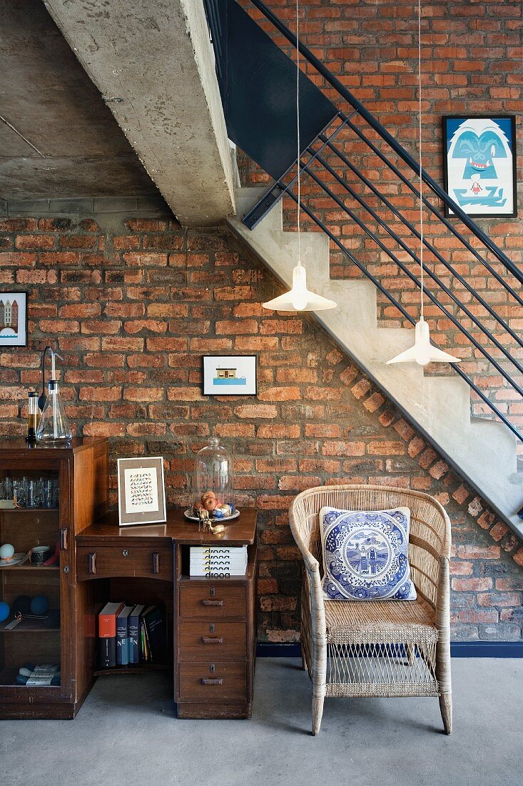 Vintage Sekretär mit kleinem Schrank und Rattanstuhl vor Treppenaufgang an Ziegelwand in loftartigem Wohnraum