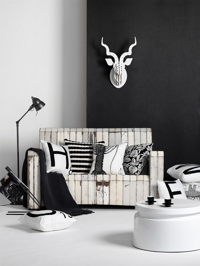Schwarz weiss Möblierung im Vintagestil - Weisser Kaffeetisch vor Holzbank mit abblätternder Farbe an schwarzer Wand und aufgehängter Tiertrophäe aus weißem Papier
