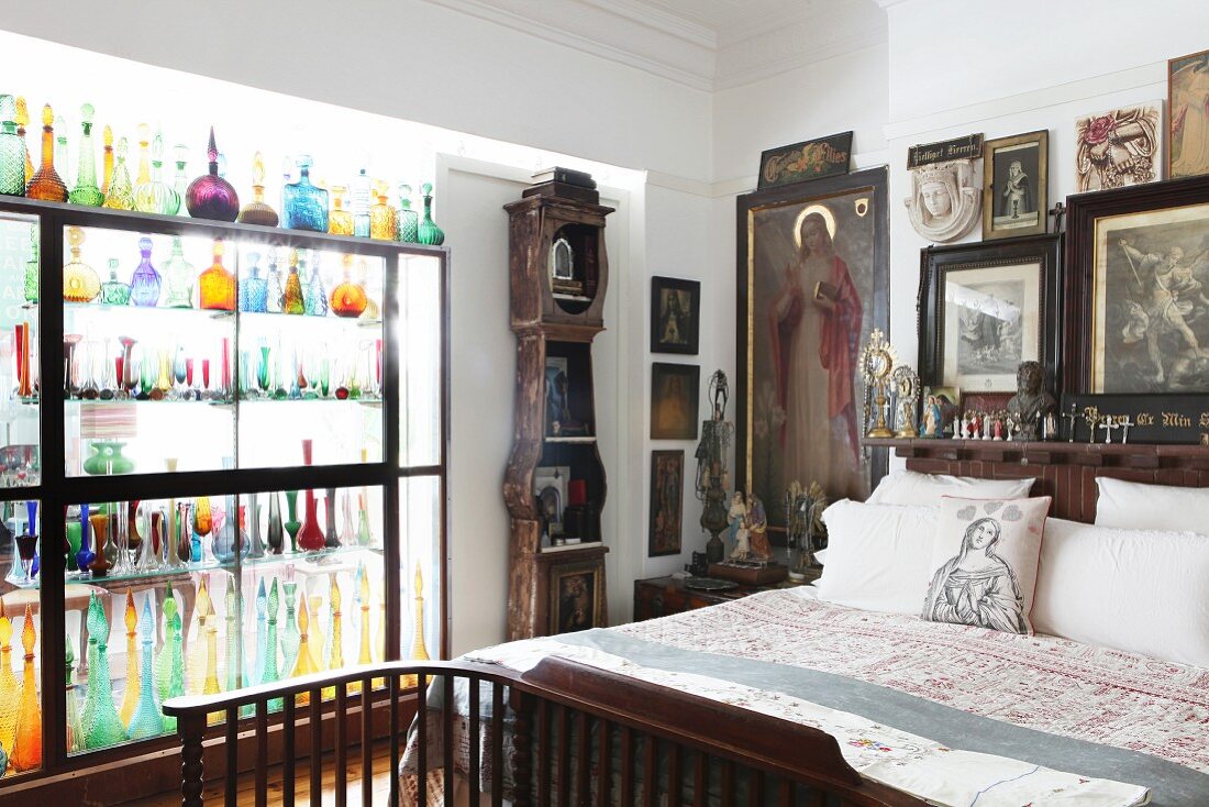 Religious pictures and statues around head of bed and coloured glass bottles in glass partition