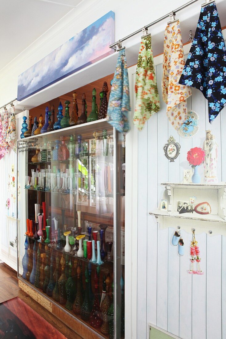 Sammlung farbiger Glasflaschen in Raumteilerregal zwischen aufgehängten, gemusterten Tüchern und Kitschgegenständen