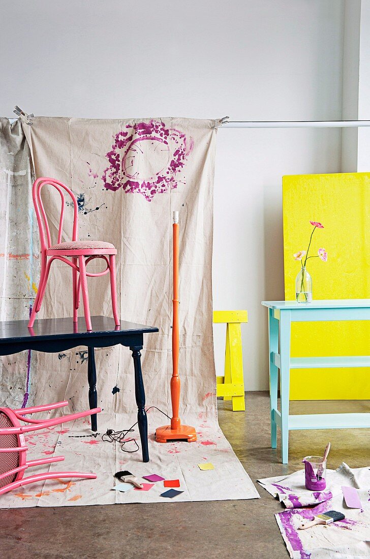 Freshly-painted wooden furniture in pink, mint-green and dark blue in front of draped dust sheets; table top and trestle painted neon yellow in background
