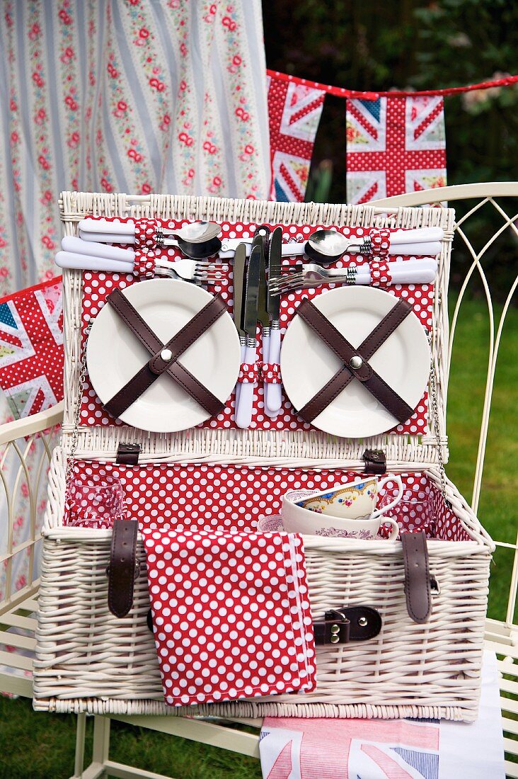 Teller und Besteck am Deckel eines geöffneten, weissen Picknickkorbes mit rot-weiss-getupftem Einlagestoff und stilisierte Union-Jack-Wimpel im Hintergrund
